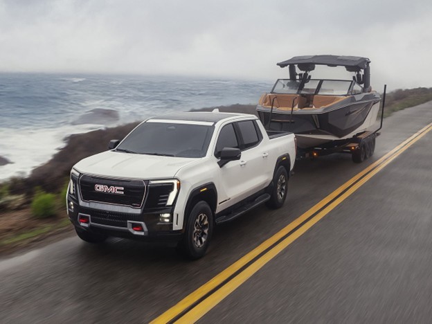 2026 GMC Sierra EV pickup truck in white towing a large boat on a coastal highway.