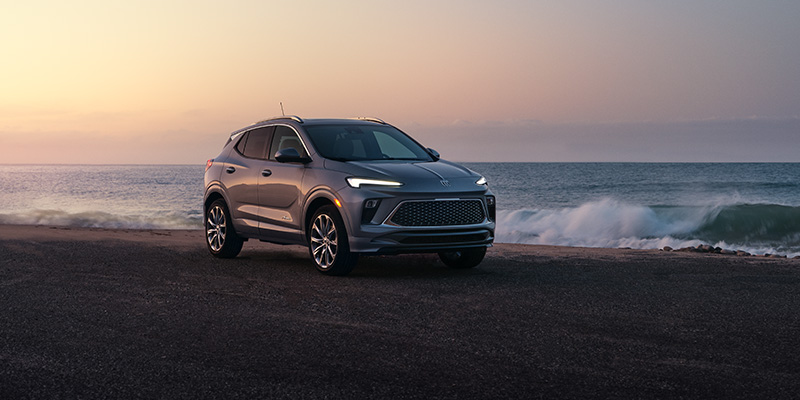 A silver Encore-GX is parked on a beach road at sunset. The sky is softly colored with warm hues, and gentle waves roll onto the shore, creating a serene mood.