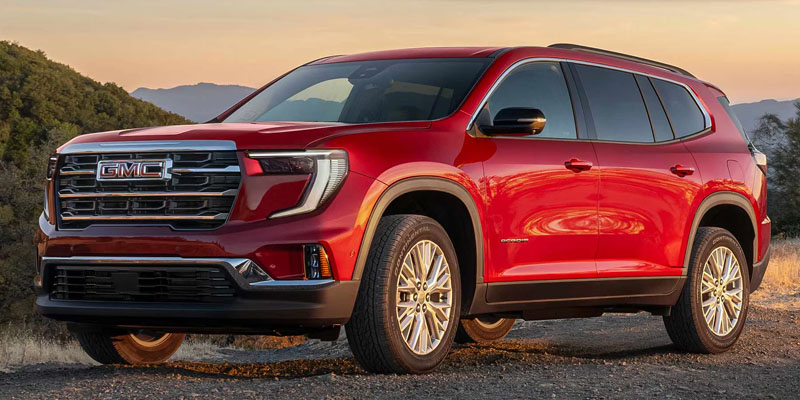 Red GMC Acadia Denali parked on a dirt road, lit by warm sunset glow. Features chrome accents, sleek lines, and large wheels. Mountains and trees in the background.