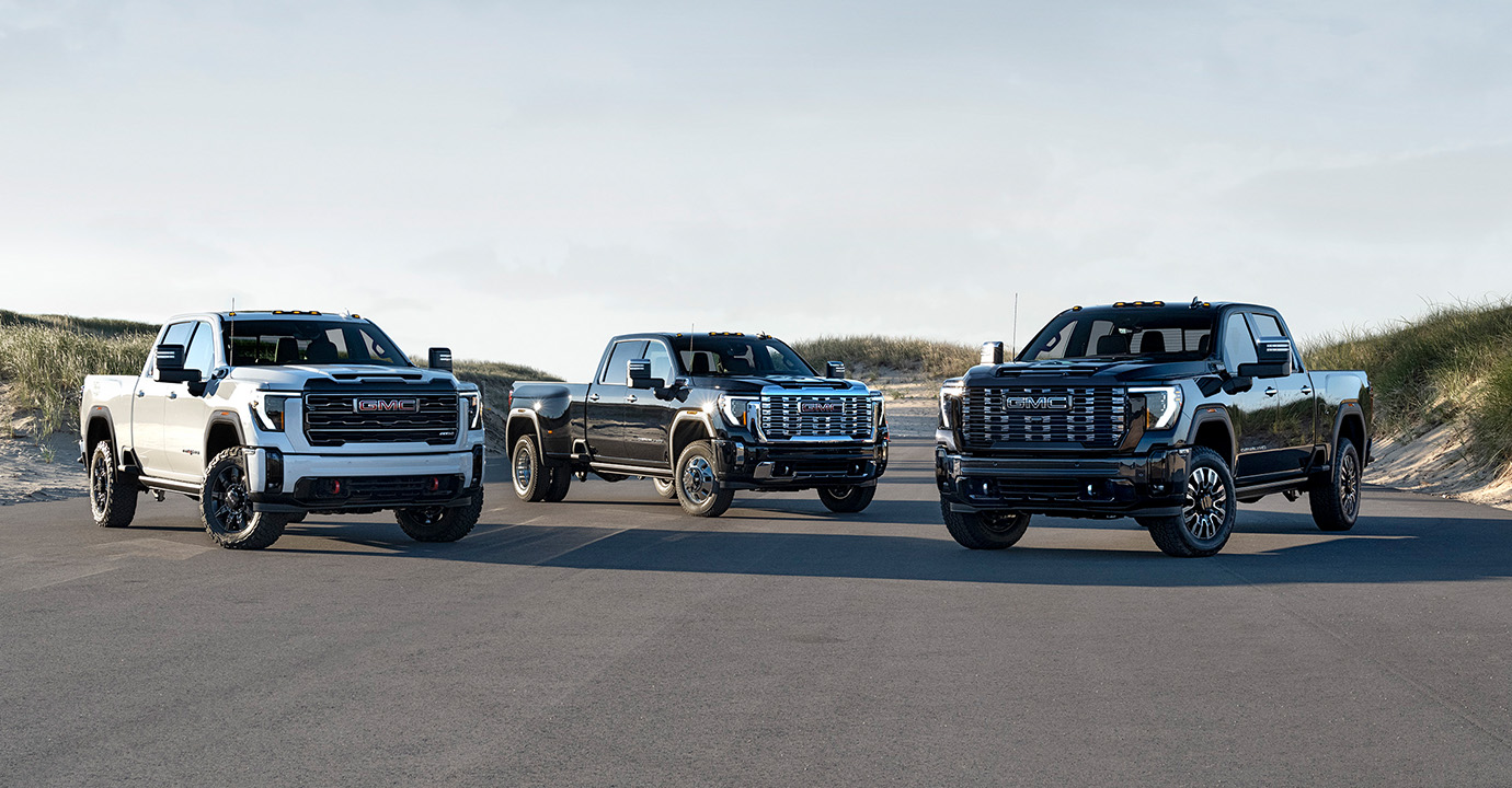 Three 2026 GMC Sierra HD trucks parked side by side on a road with grassland in the background.